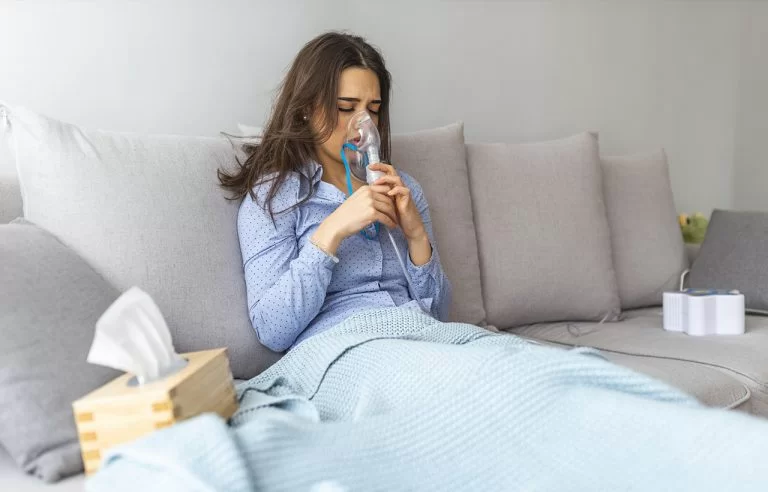 Mulher deitada na cama toda coberta fazendo nebulização e com lenços de papel perto dela